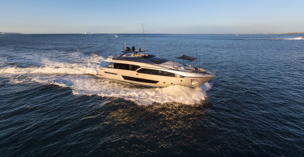 A sleek, champagne-colored motor yacht cruising across the ocean at sunset, creating a large white wake.