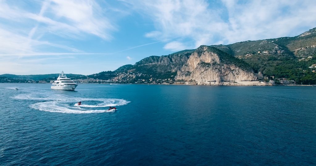 An aerial wide shot of a large white multi-deck luxury yacht and two jet skis cruising through deep blue water. The jet skis leave circular white wakes in the foreground, while a rugged, mountainous coastline with greenery and sheer cliffs rises in the background under a bright, blue sky.