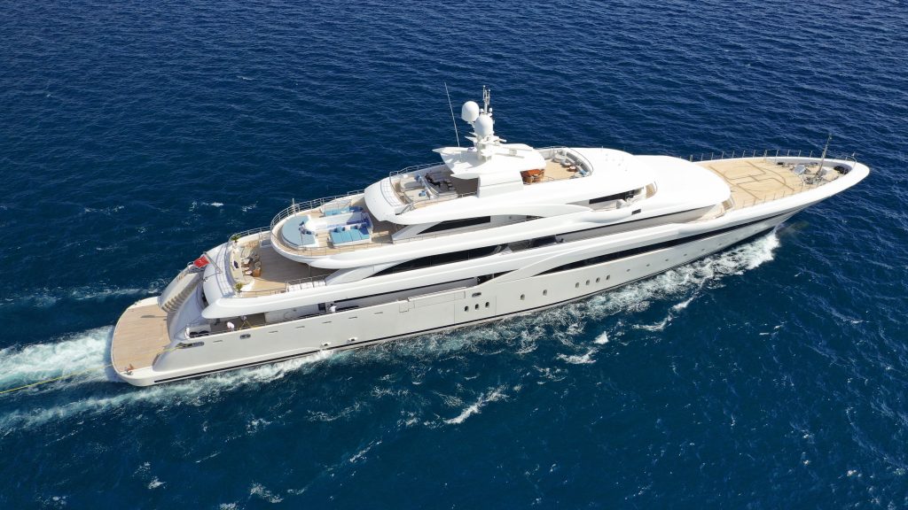 High-angle aerial view of a large white multi-deck superyacht cruising through deep blue water, showing lounge areas with blue cushions and a helipad on the bow.
